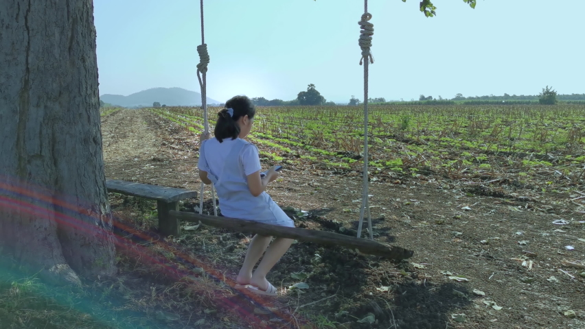 Little Girl in gray jeans overalls play or watch a smart mobile phone on the swing under the shade of a large tree in the sunflower sprout garden with lens flare.