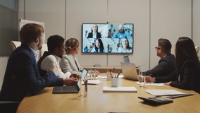Dolly-in shot of team of businesspeople sitting at table in meeting room and discussing work with their colleagues via video call
