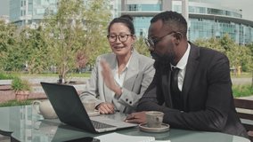 Medium shot of Asian businesswoman and African-American businessman in formal wear sitting at table in outdoor cafe and having conversation with someone on video call on laptop