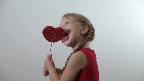A girl in a red dress stands in profile, holds a lollipop - her heart and licks, nibbles on caramel candy. The child is eating sweet candy. Enjoy dessert. Concept - 14.02, Valentine's Day