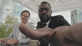 POV shot of African-American businessman and Asian businesswoman in formal wear sitting at table in outdoor cafe in city and talking on video call while looking at camera