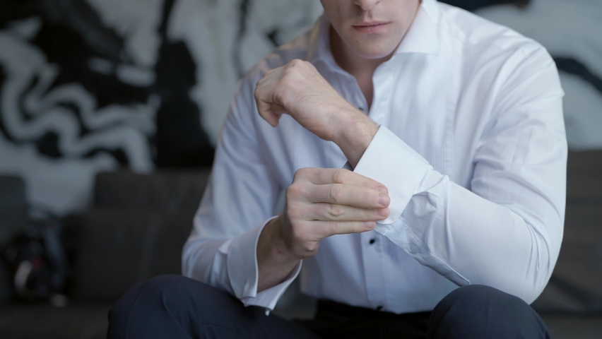 The man gets dressed and does his buttons on the sleeves of his white shirt. Action. Close up of male arms, man sitting and trying to button up.