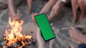 Close-up shot of green screen template smartphone in female hands on the beach by the fire, girl is watcing content without touching gadget screen. Modent technology and information concept.