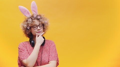Caucasian Woman Wearing Bunny Ears Hairband And Spectacles With Headphones Around Her Neck Pondering Thoughts In Yellow Background. medium shot