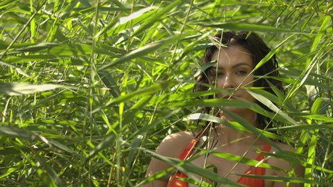 A young woman in the bulrush looks at the camera. Natural surroundings outside. The woman's face is lit by the sun's rays in the tall grass. Slowmotionie.