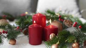Christmas decoration with red candles, spruce branches, cones, balls and garland on white table on blurry background. Side view. Hand of woman puts one candle. New year mood, festive concept