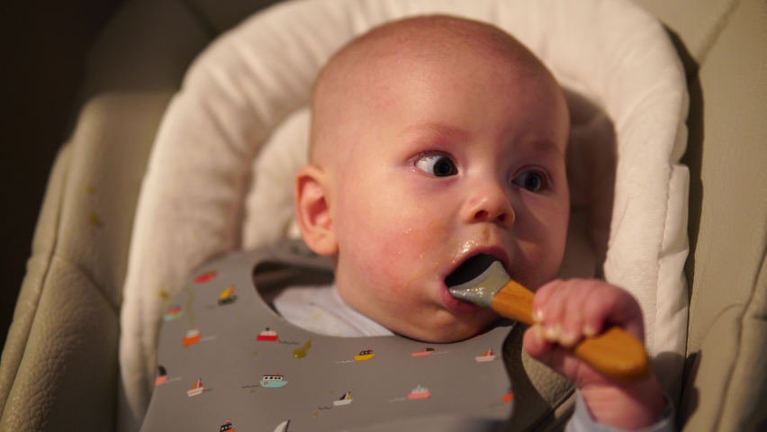 Baby boy sitting on feeding chair gnawing rubber spoon. Cute infant teething and biting spoon after eating.