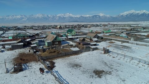 Bird’s eye view Buryatia winter Arshan rural settlement, authentic wooden houses among high snow-capped mountains. Epic suburban rural landscape. Baikal region Siberia. Typical Russia ancient village