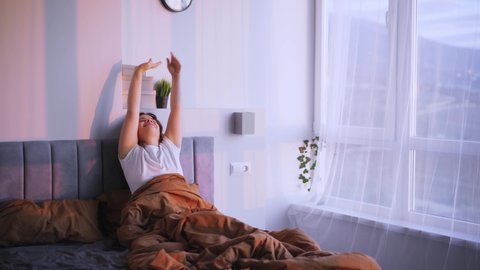 Cheerful young woman enjoying early morning awaking. Smiling happy lady rubbing sleepy eyes, stretches and rising up arms while waking up in large bed 