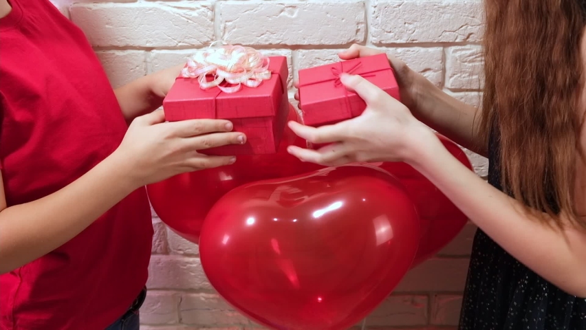 Little boy and girl gives Valentines Day red Gift Boxes. Heart shapes balloons background. Happy smiling kids celebrating Valentine