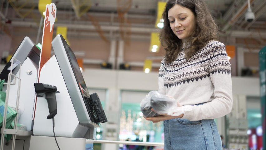 A young woman at a self-service checkout in a grocery supermarket makes purchases, punches barcodes on the product and pays Royalty-Free Stock Footage #1084316626