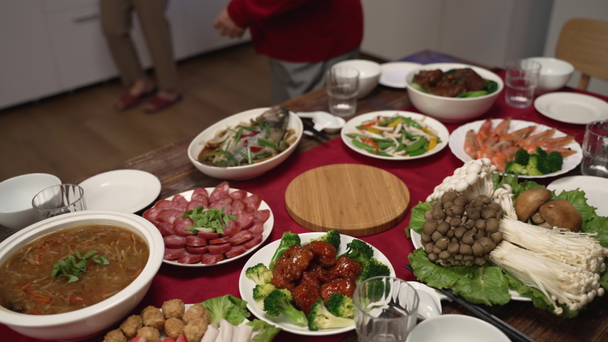 happy asian senior woman grandmother wearing red clothes looking at daughter in law carrying soup on dinner table. getting ready for family reunion meal at home. word on cabinet translation: fortune