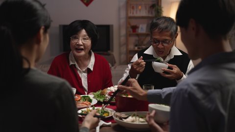 selective focus with over shoulder view smiling asian elderly couple grandfather and grandmother holding food with chopsticks and giving to their daughter in law at dinner table on eve of new year