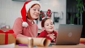 Online Xmas party. Close up portrait of cheerful mother and daughter wearing funny holiday headwears waving hands to laptop, video chatting with family on distance, tracking shot, slow motion
