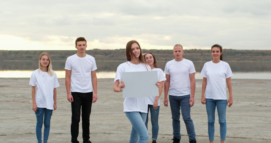 A girl in a white T-shirt holds a square gray sheet of cardboard with her hands in front of her and pushes it forward, in the background young people show their thumbs up.