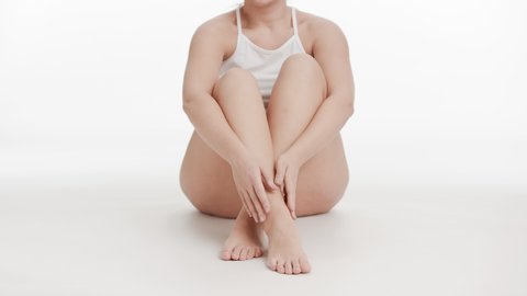 Horizontal medium long shot of white-skinned good-looking plus size woman in white top strokes her crossed legs sitting on the floor on white background | After leg shaving concept