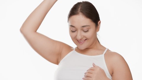 Pretty Caucasian young plus size woman with long dark hair in ponytail in white top strokes her arm and armpit smiling wide for the camera on white background | Smooth armpit concept