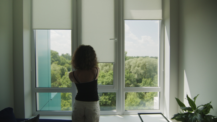 Rear view of young girl with curly hair that pulls up fabric blinds on a window. Sunny summer day Royalty-Free Stock Footage #1084325650