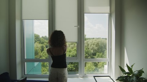 Rear view of young girl with curly hair that pulls up fabric blinds on a window. Sunny summer day