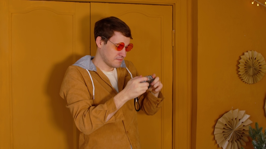Young man in orange glasses with old photo camera stands against background of brown doors in room. Stylish male considers old camera and takes photo, looking at camera.