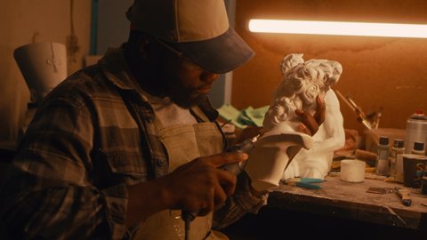 African American craftsman in checkered shirt and cap polishing half of plaster bust while sitting near illuminated workbench and working in sculpting workshop at night