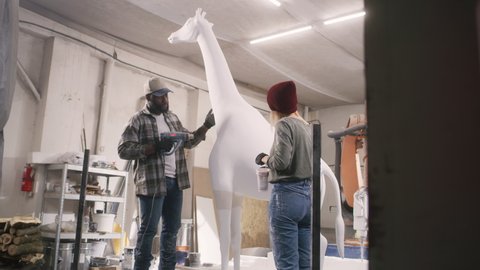 Pan left view of woman mixing paint in cup and checking statue while black man drying white pigment on giraffe near robotic arm milling machine during work in spacious workshop