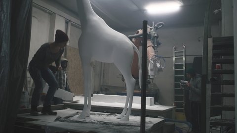 Group of diverse artisans turning on lights and entering workroom then starting to work with gypsum giraffe statue and polymer blocks together