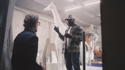 Low angle of mature customer in suit discussing gypsum giraffe statue with black craftsman in illuminated workroom