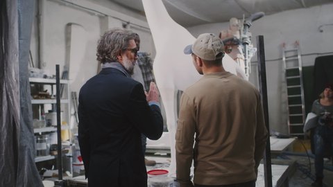 Zoom in view of male client and artisan talking and shaking hands while watching employees crafting giraffe sculpture in workshop