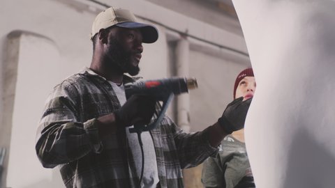 Low angle of woman speaking with African American colleague with dryer while painting giraffe statue in professional workshop together