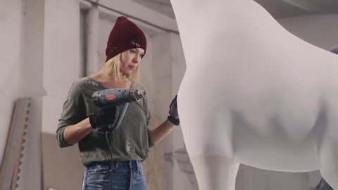 Female artisan in casual clothes and hat drying white paint on plaster giraffe sculpture while working in professional creative workshop