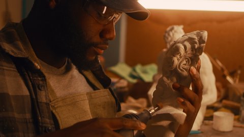 African American craftsman in checkered shirt and cap polishing half of plaster bust while sitting near illuminated workbench and working in sculpting workshop at night