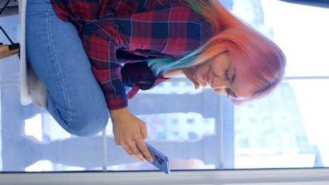 Young girl with dyed hair using mobile phone. Vertical stock video of female browsing news feed on social media app in modern smartphone. Royalty free 4K footage of young woman using cellphone