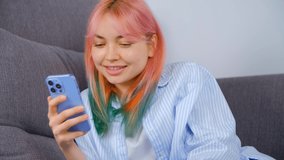 Happy girl with dyed hair browsing internet on mobile phone. Individual young woman with colored hair using app on smartphone while lying on couch at home. Royalty free 4K video of smiling female