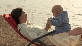 Happy mother plays with son against sun light glittering on water at sunset, mom with baby shining in backlighting. High quality 4k footage