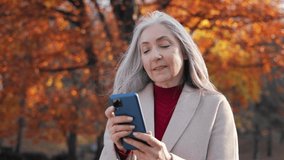 Active senior people with digital technology concept.Smiling elderly gray-haired woman using smartphone for texting message,reading news,watching photo or video in social media walking in autumn park.