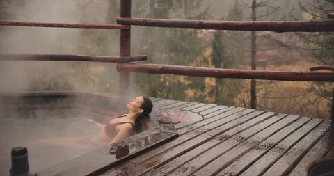 Young woman in a bikini bathing in wooden bath outdoors while raining. Mountain resort spa and entertainment