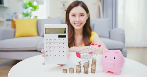 asian woman woman shows calculator with coins and piggy bank at home happily