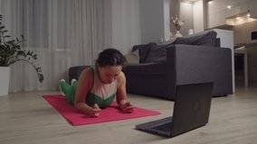 Adult woman in sportswear performing abs workout, doing yoga plank exercise, watching online video tutorial on laptop, getting exhausted, falling on mat floor indoors. Training for weight loss at home