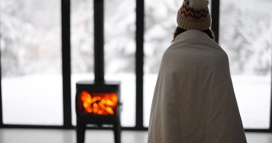Woman enjoys winter time at home with burning fireplace and panoramic window, looking outside on snowy landscape. Concept of winter mood and comfort at home. Idea of winter vacation in the mountains