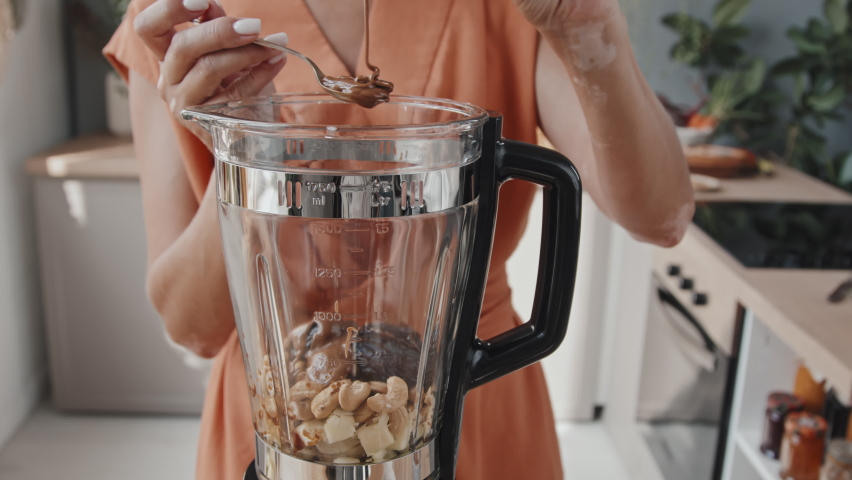 Handheld close up of unrecognizable young woman adding buckwheat honey to figs and cashews in blender while making smoothie in morning
