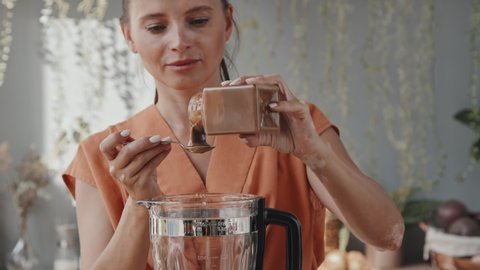 Tilt down shot of young vegetarian woman adding spoonfuls of tasty buckwheat honey to cashews and figs in blender while preparing smoothie in kitchen