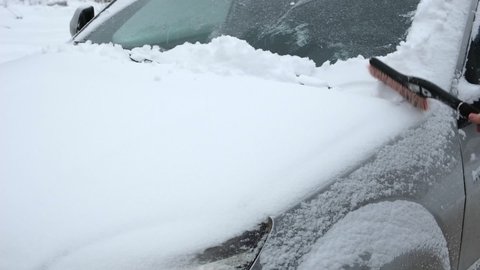 Person brushing fresh snow from car windows after snow storm blizzard. Morning routine difficulties of car owners in cold winter weather. Car window cleaning. Winter season. White snow. Vehicle care.