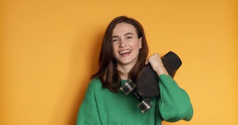 Stylish young happy girl in green sweater smiling and showing tongue with skateboard. Showing shaka sign in front of yellow wall. Enjoying life. Healthy and careless lifestyle.