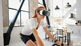 Young red-haired woman in sportswear and VR headset is pedaling indoor cycle trainer. Studio apartment, kitchen. Sunny day. Daily training at home. Modern technologies. Close up, slow motion