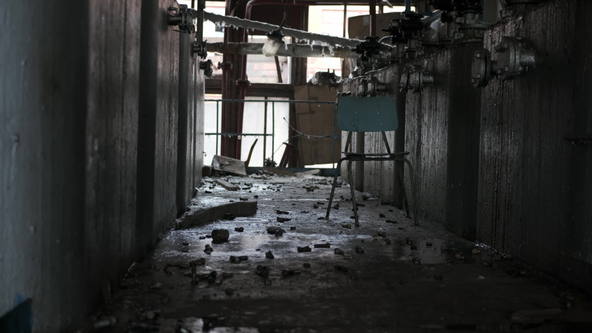 Aisle between equipment or barrels of chemical liquid in old abandoned factory, in middle there is old wooden chair, concept of loneliness in big world. In background, sun shines through large window.
