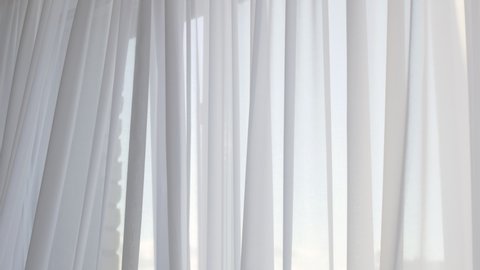 The frosted white curtain of the window blazes with a gust of wind. The curtain inflates as much as it fills the entire room. The curtain develops in the wind by the window.