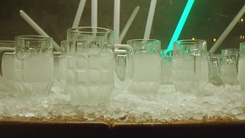 Row of many empty frozen 0.3 l and 0.5 l beer glasses . Camera moving across beer cups .  Neon green flashing lights  . Ice cubes on table . Slow motion . Shot on ARRI ALEXA Camera with Laowa lens . 