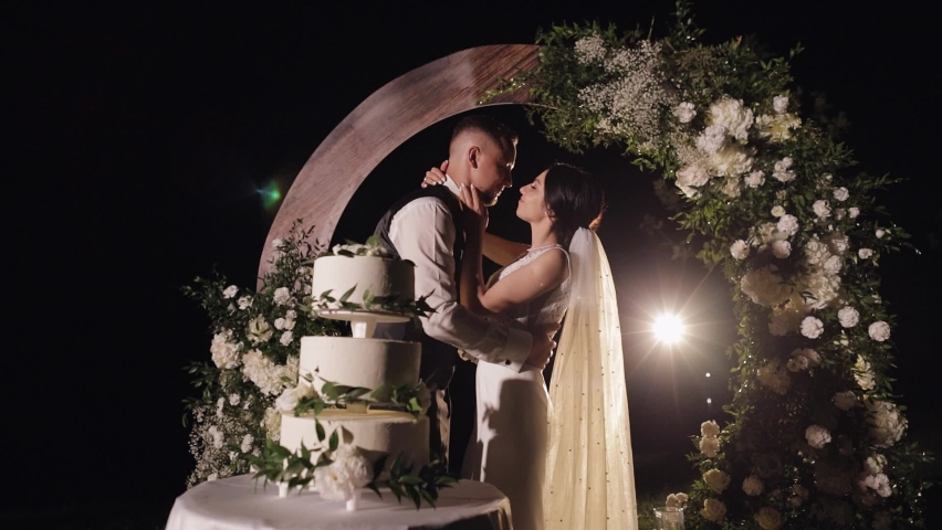 Newlyweds family making a kiss after cut of wedding cake. Lovely bride and groom couple standing near wedding cake dessert. Lovely man and woman with three-tiered wedding cake. Outdoors, slow motion Royalty-Free Stock Footage #1084407157