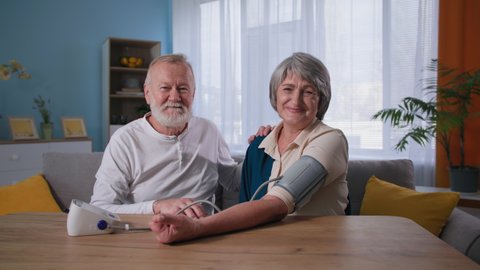 elderly happy couple looking at camera while taking blood pressure, husband using blood pressure monitor for wife with hypertension at home
